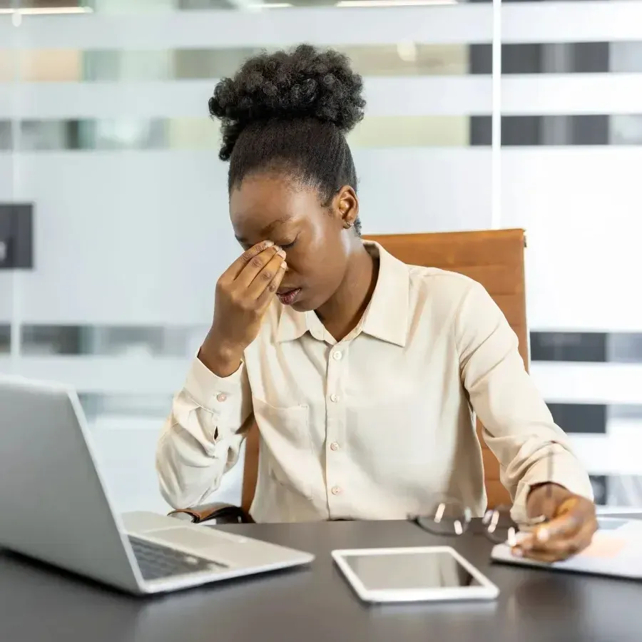 Person at desk rubbing eyes beside laptop and tablet, looking tired and stressed