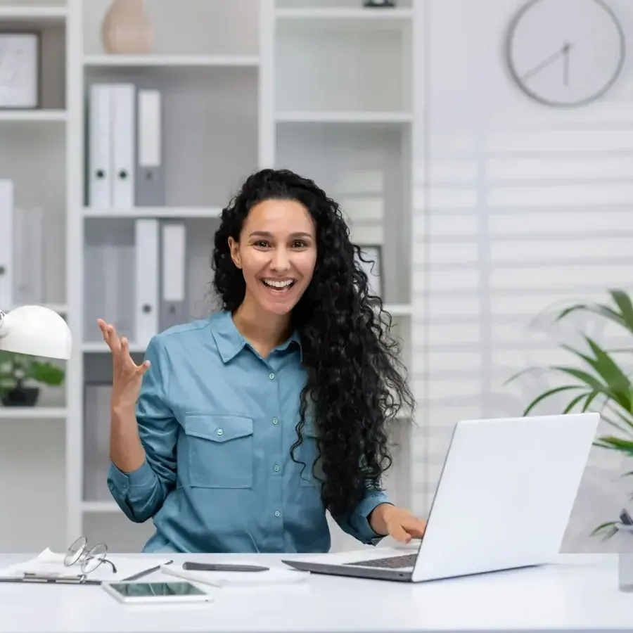 Smiling woman in a blue shirt standing at a desk with a laptop in a bright office