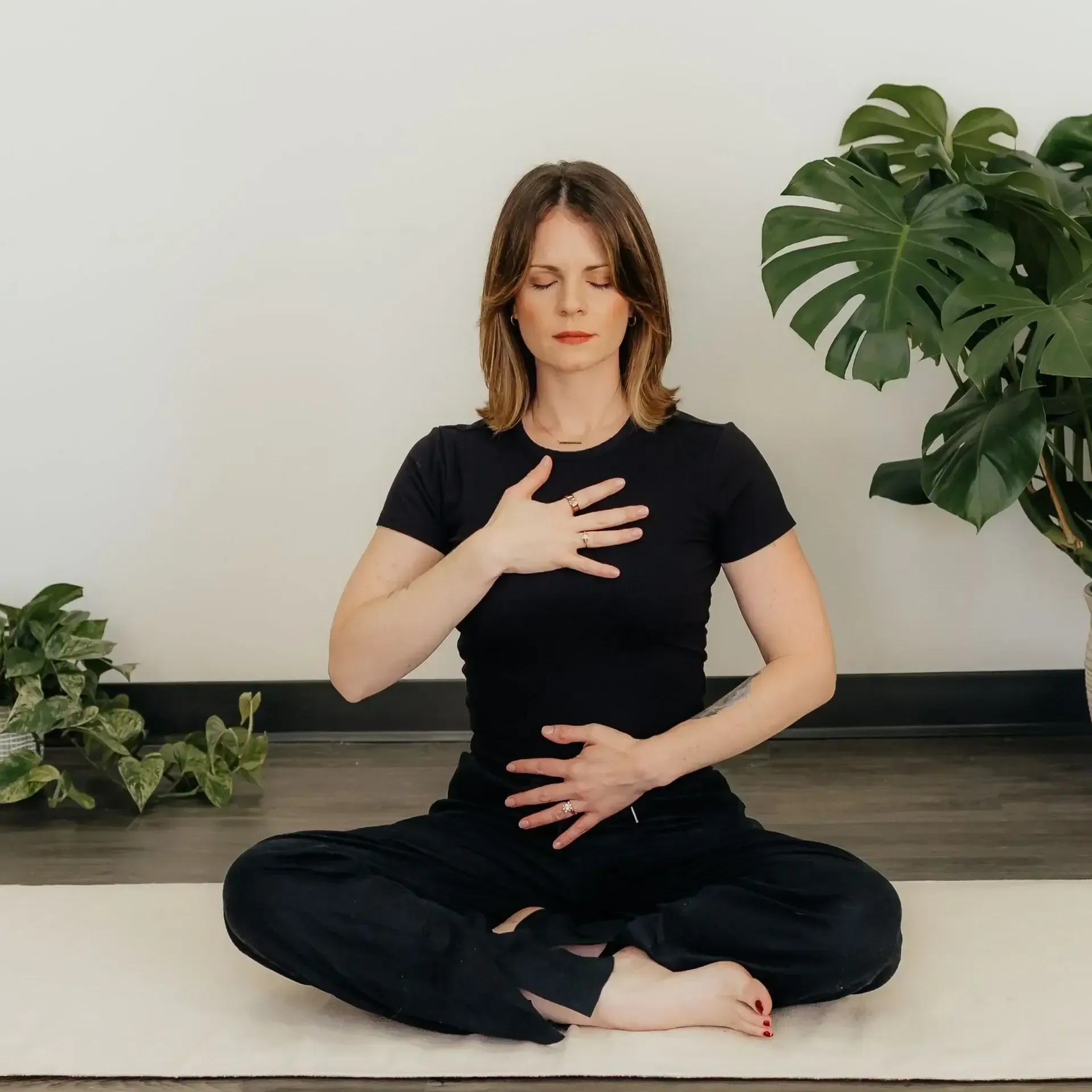 Person meditating cross-legged indoors, hands on chest and abdomen, with a plant in the background