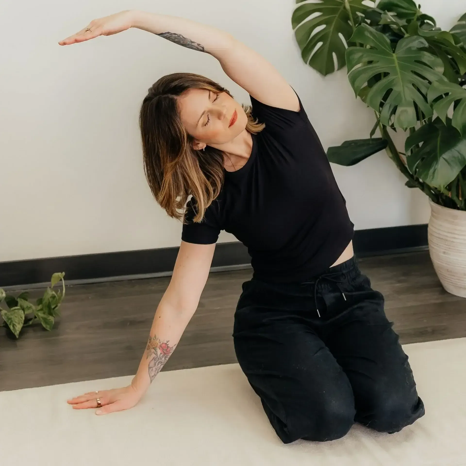 Person kneeling and side-bending in a yoga pose indoors beside a potted plant