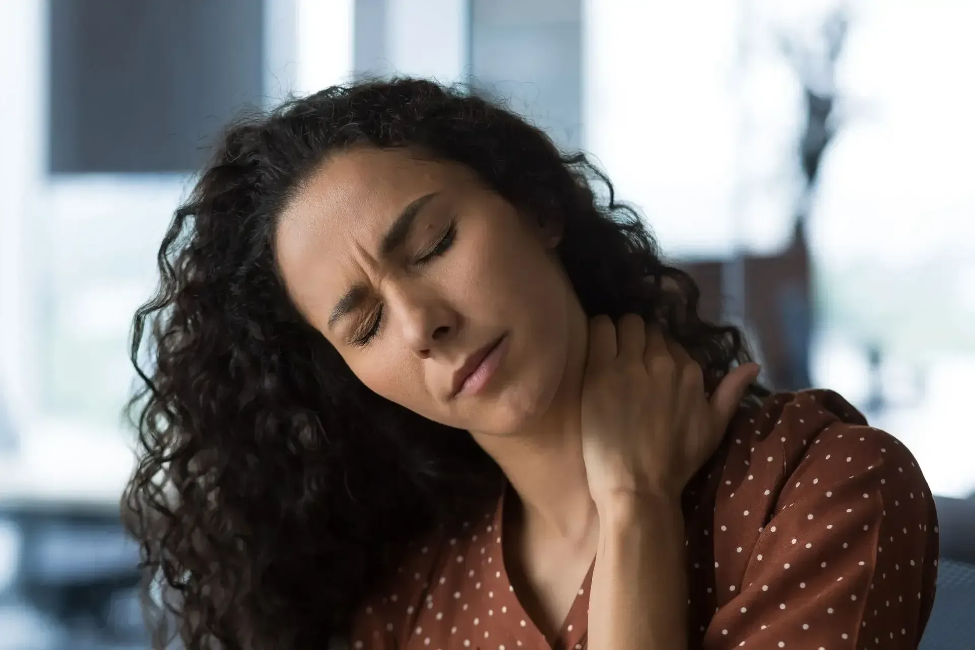 Woman with curly hair holding her neck and wincing, seated indoors near a window.
