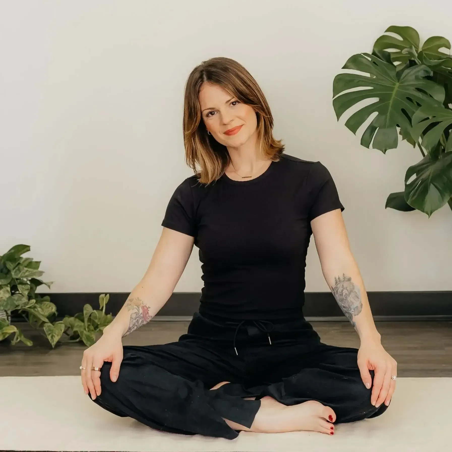 Person sitting cross-legged on a floor in a black outfit, in a bright room with plants.