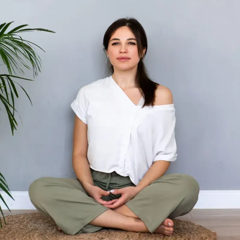 Woman sitting cross-legged on a mat in a bright room, wearing a white top and olive pants, with a plant nearby