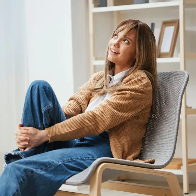 Woman sitting in a chair indoors, hugging her knee and looking up thoughtfully