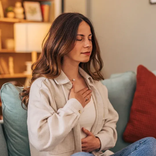 Woman seated on a couch, hand on chest, eyes closed, in a cozy living room.