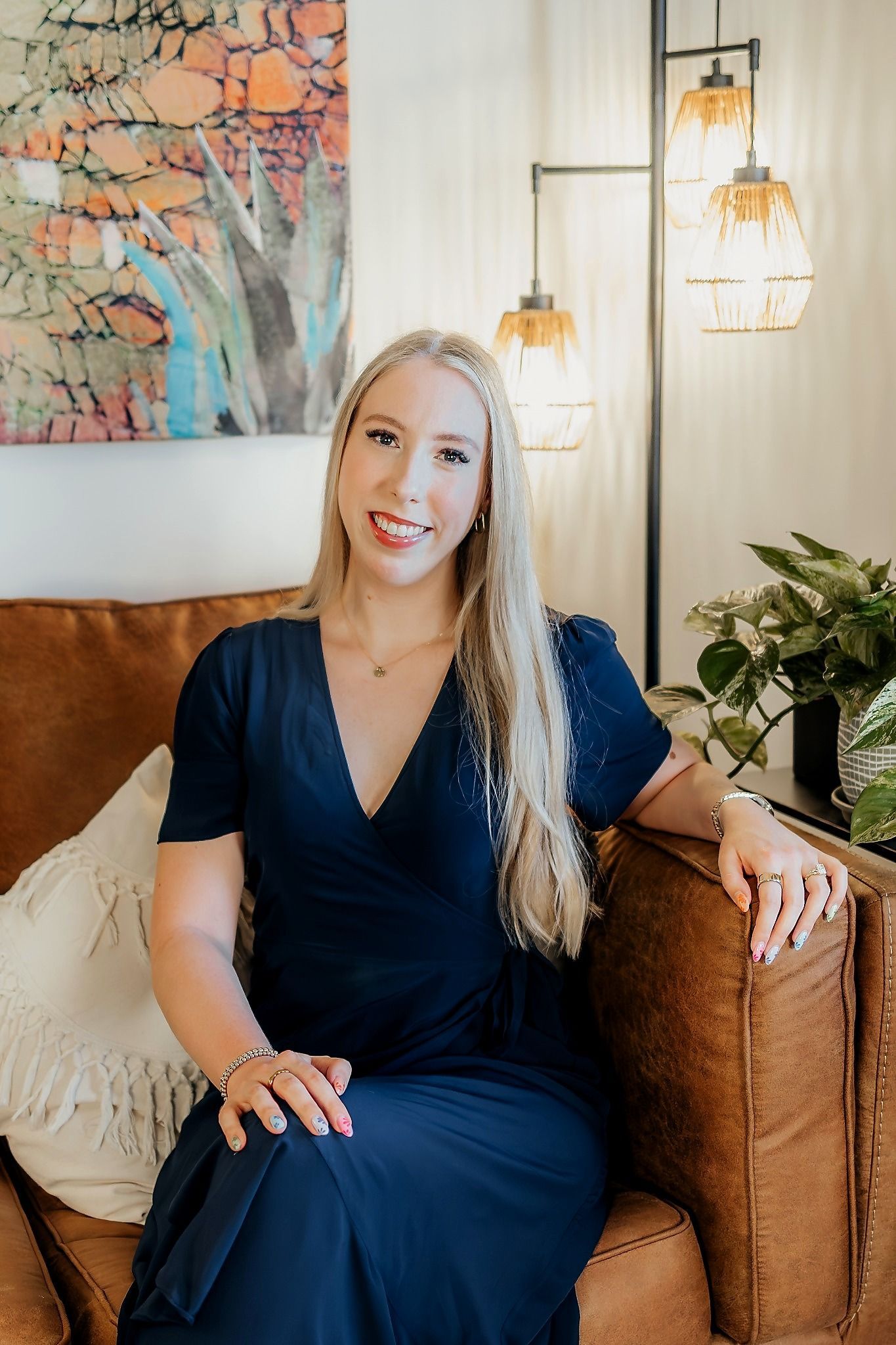 Woman in a navy dress seated on a brown sofa in a warmly lit living room, smiling beside a plant and wall art