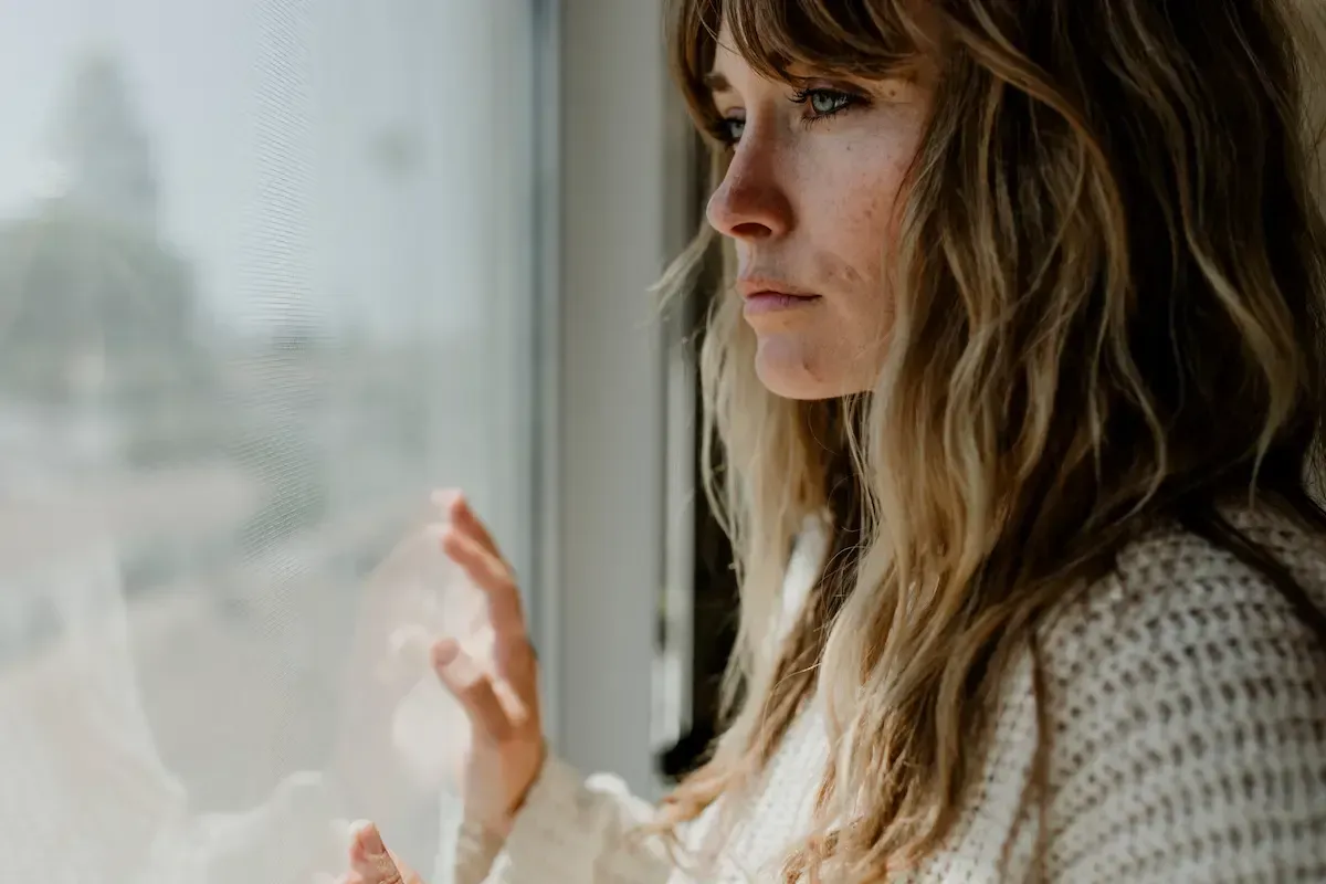 Woman in profile by a window, gazing out with a pensive expression, wearing a cream sweater.