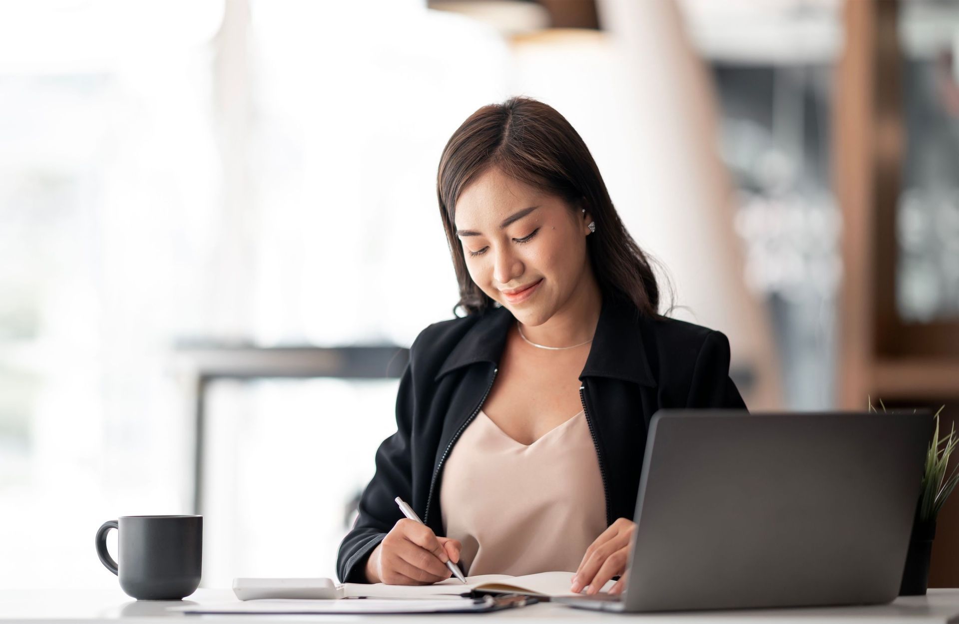 Woman working an administrative job at desk writing in notebook with coffee mug and laptop in front of her