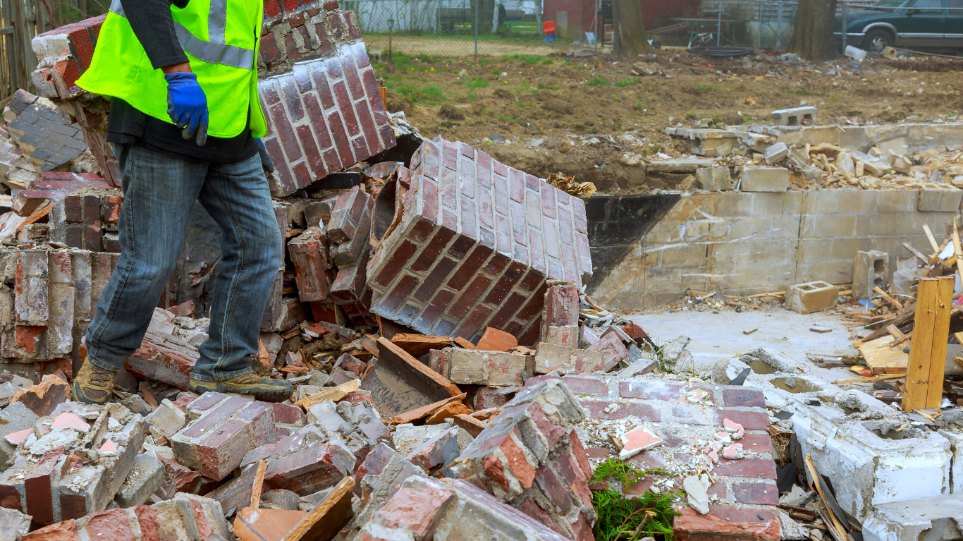 A worker in a high-visibility vest stands amidst a pile of red bricks and debris at a construction demolition site.