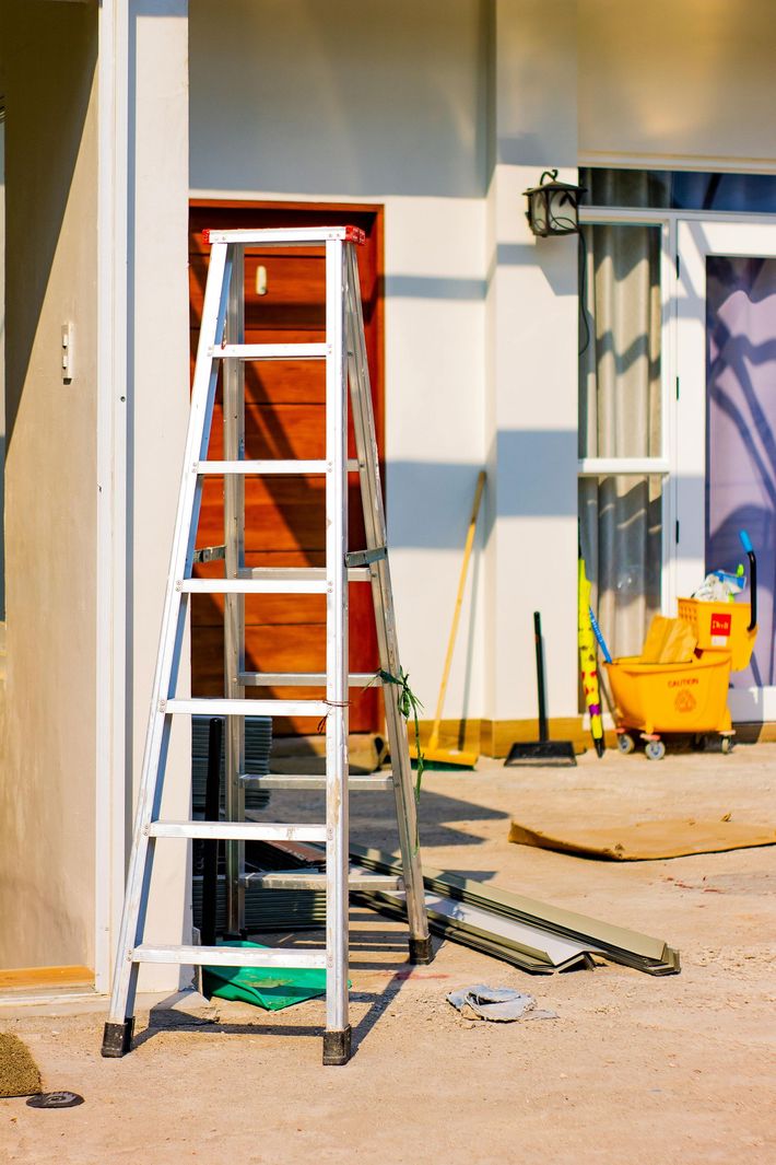 A metal A-frame ladder stands on a patio next to a building entrance with a yellow mop bucket and cleaning tools nearby.