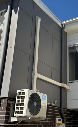 Exterior wall with gray panels, exposed white piping, brick base, and a mounted air conditioner unit.