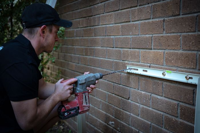 Technician kneeling beside an open outdoor electrical panel, inspecting wires next to a house.