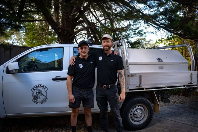 Two workers in black uniforms standing beside a white utility truck under a tree
