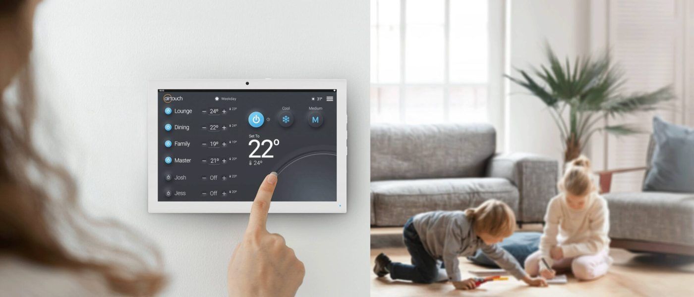 Hand holding a tablet displaying weather in a bright living room with children playing on the floor.