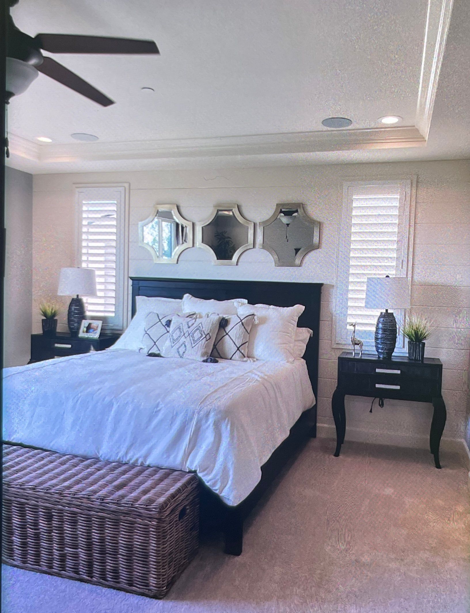 Bedroom with bed, nightstands, decorative mirrors, shutters, and ceiling fan. Light-colored walls and carpet.