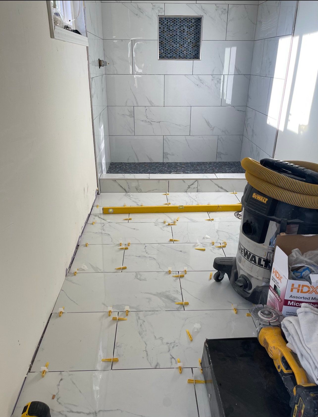 Bathroom under construction, white marble tiles, black shower floor. Tools and level visible.