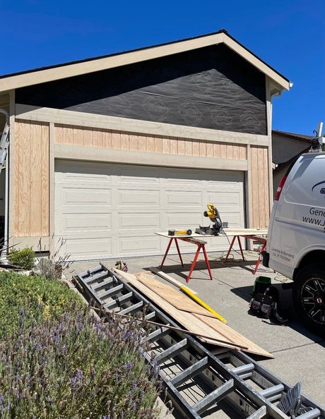 Garage exterior under construction; plywood siding, black roof trim, power saw, ladder.