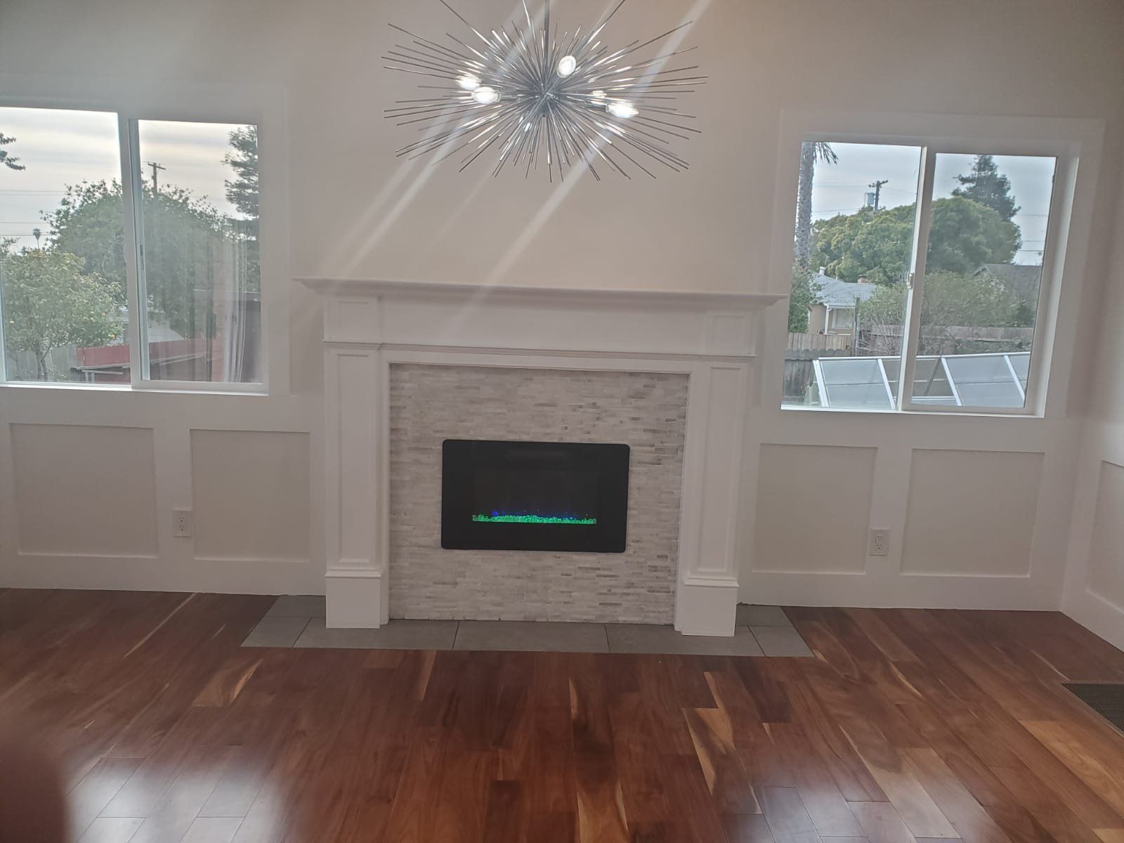 Interior view: Fireplace with white mantle between two windows. Hardwood floor. Chandelier.