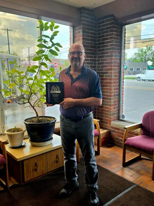 Man holding a plaque beside a potted tree in a brick-walled room with large windows and a chair