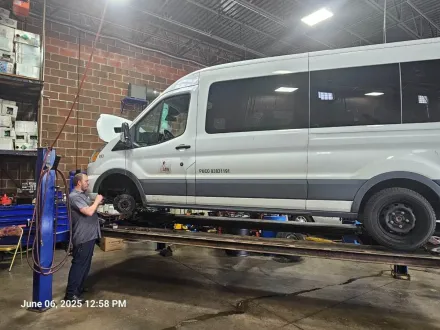 White passenger van on a hydraulic lift in an auto repair shop, with two workers inspecting underneath.