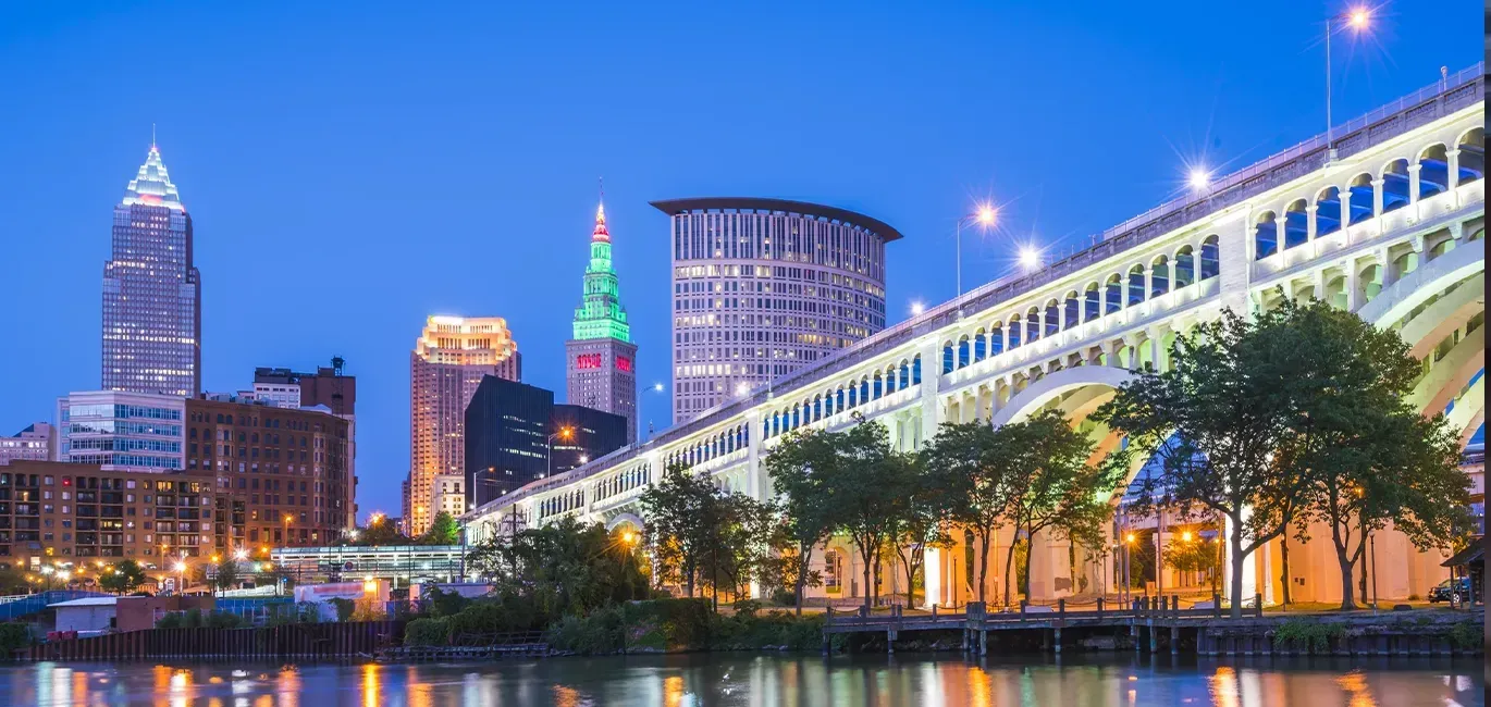 City skyline at dusk with illuminated riverwalk and a lit bridge reflecting on the water