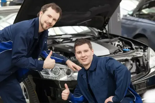 Two mechanics in blue uniforms giving thumbs up in front of an open car hood