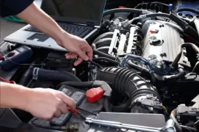 Hands working on a car engine, using tools near the battery and intake hose.