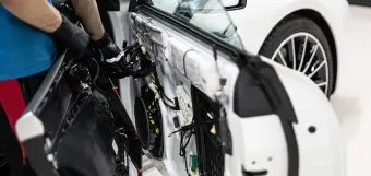 Technician repairing the inside of a white car door with tools, wiring exposed.