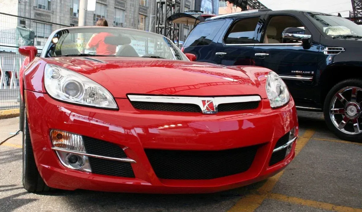 Red sports convertible parked on a city street, front view, with a black SUV beside it.