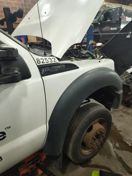 White pickup truck with hood open in a garage, front wheel removed and body damaged during repair