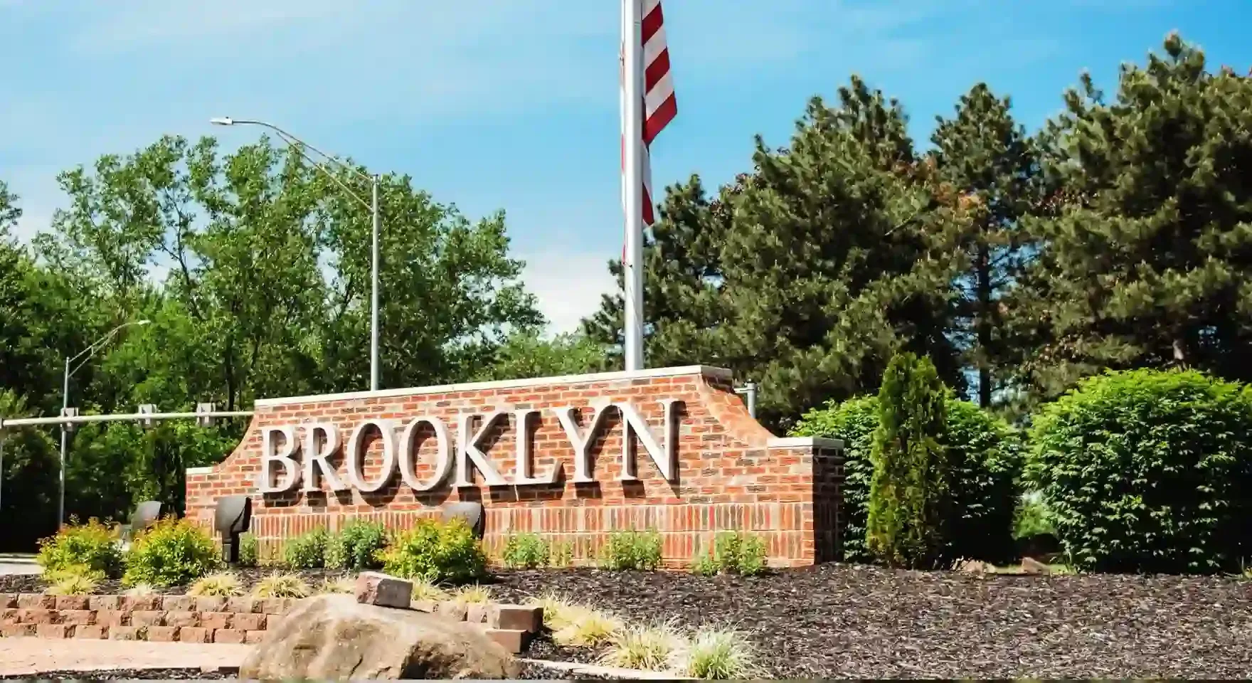 Brooklyn stone entrance sign in a landscaped area with trees and a flagpole under a blue sky