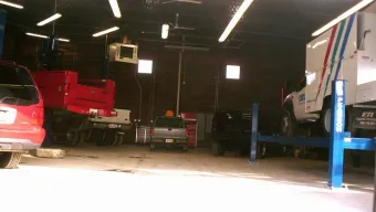 Auto repair shop with cars on lifts, including a red vehicle and a white truck inside a dim garage.