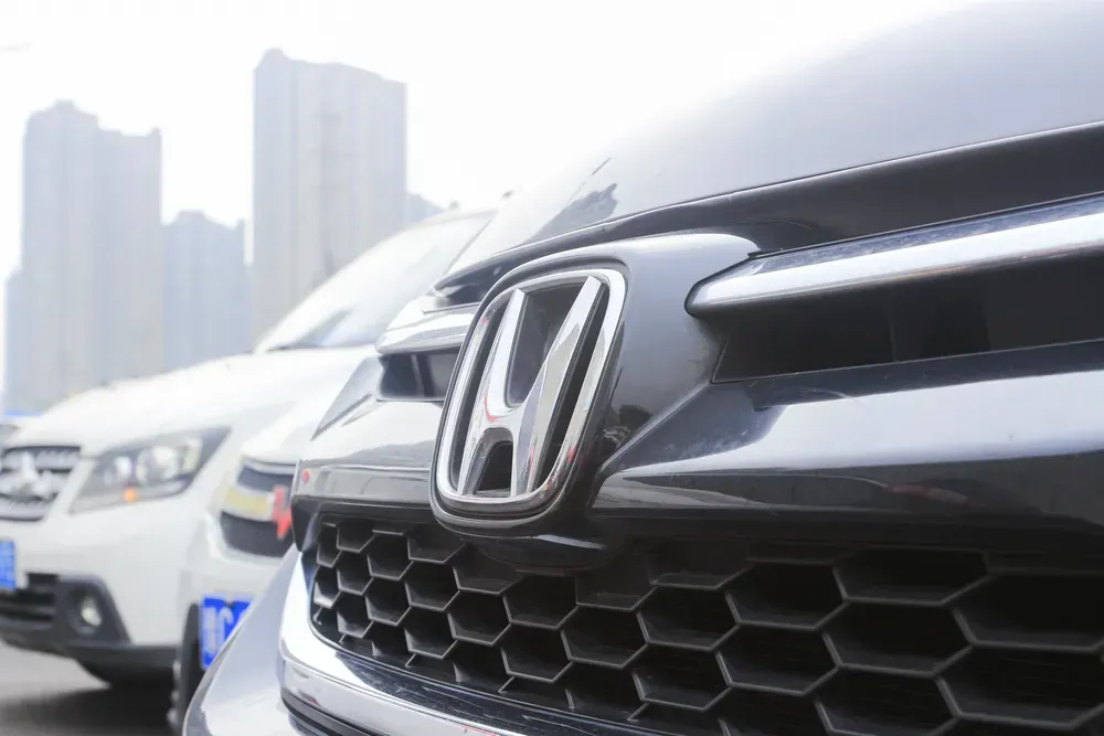 Close-up of a black Honda car grille with blurred white cars and city buildings in the background