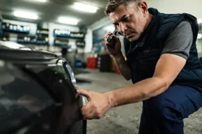 Mechanic crouching beside a car in a garage, inspecting the front bumper with a tool in hand