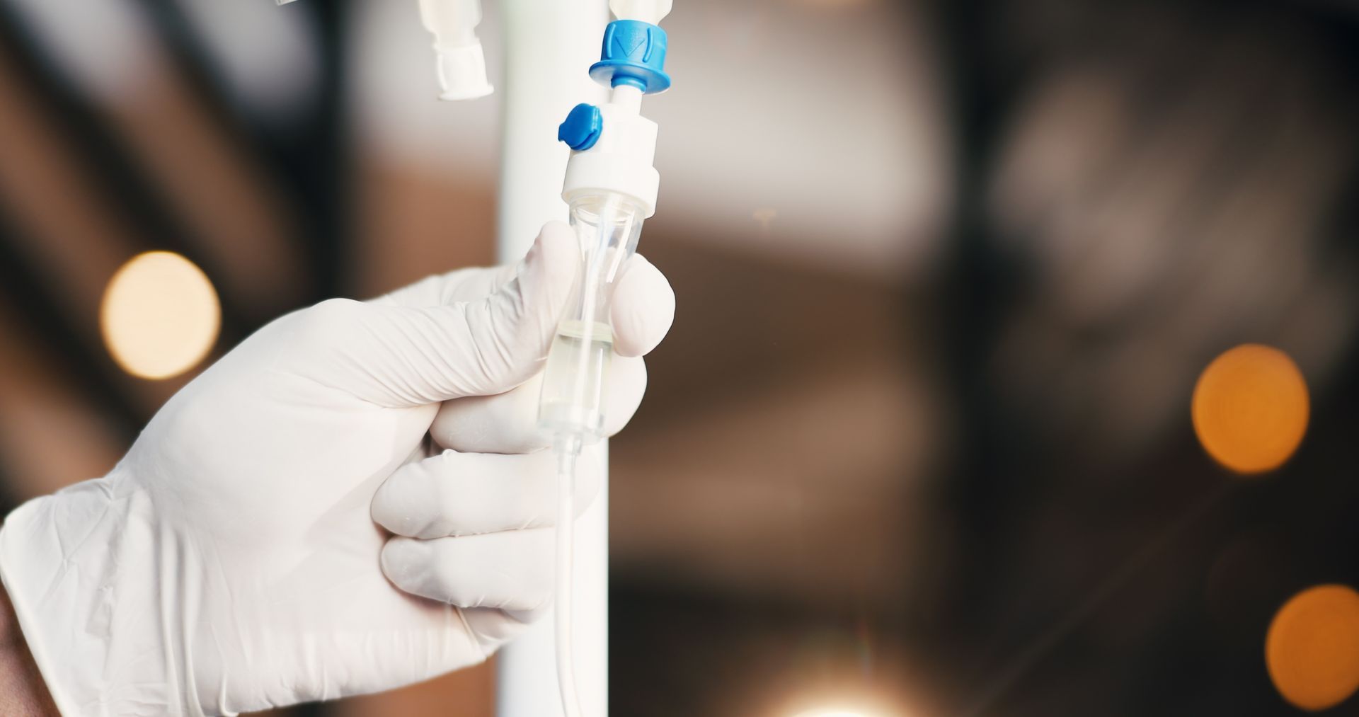 Gloved hand holding a clear IV tube with blue connector, against a blurred background