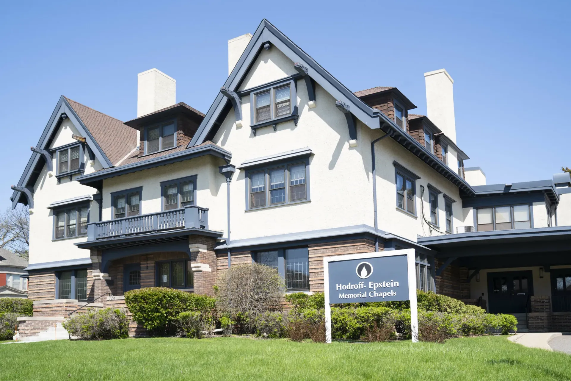 White Tudor-style house with dark trim, balconies, and a sign in front on a sunny day