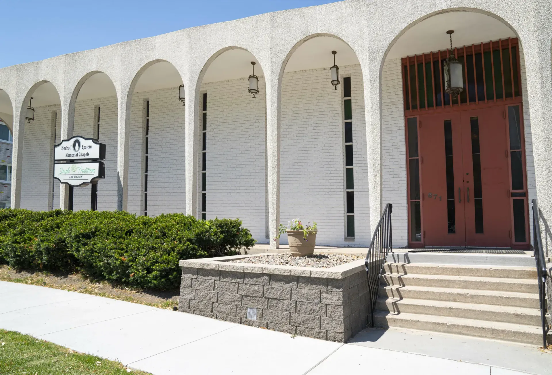 White stucco building with arched columns, red door, steps, and a hedge in front
