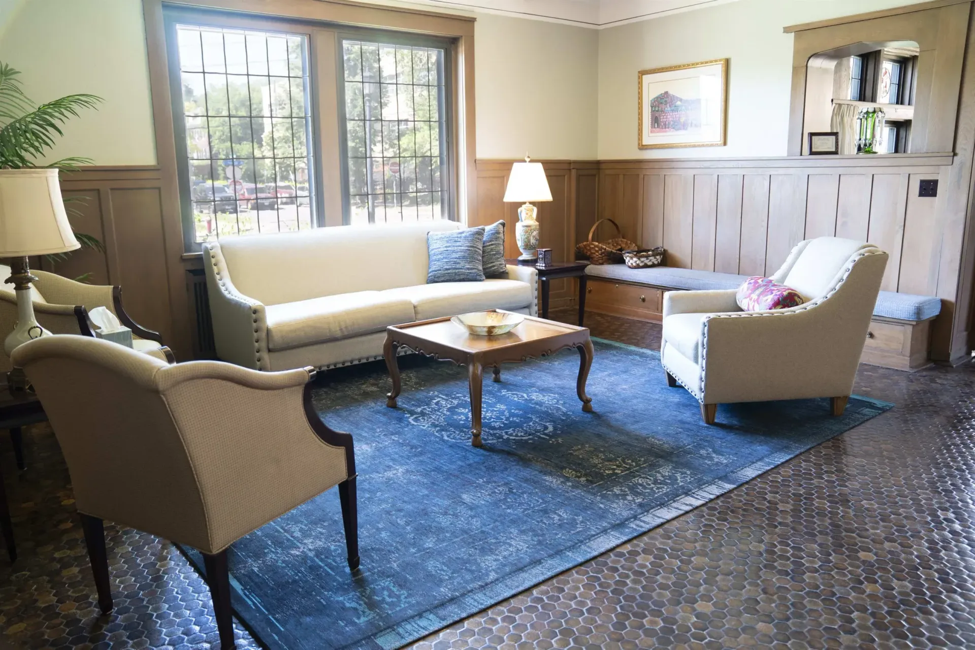 Cozy living room with cream seating, blue rug, coffee table, lamp, and large windows with daylight.