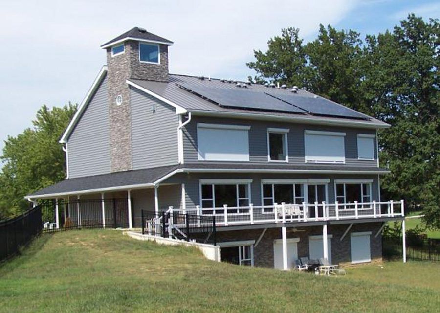 Gray two-story house with a wraparound deck on a grassy hillside, backed by trees.