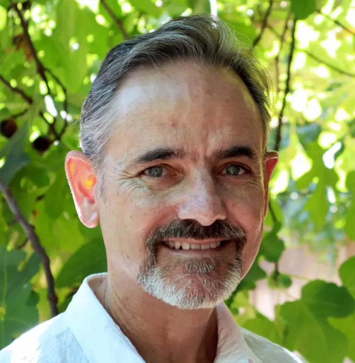 Man with salt and pepper hair and beard, smiling in front of green foliage.