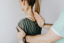 A person's back being examined by someone's hands. They are in a room with a light wall.