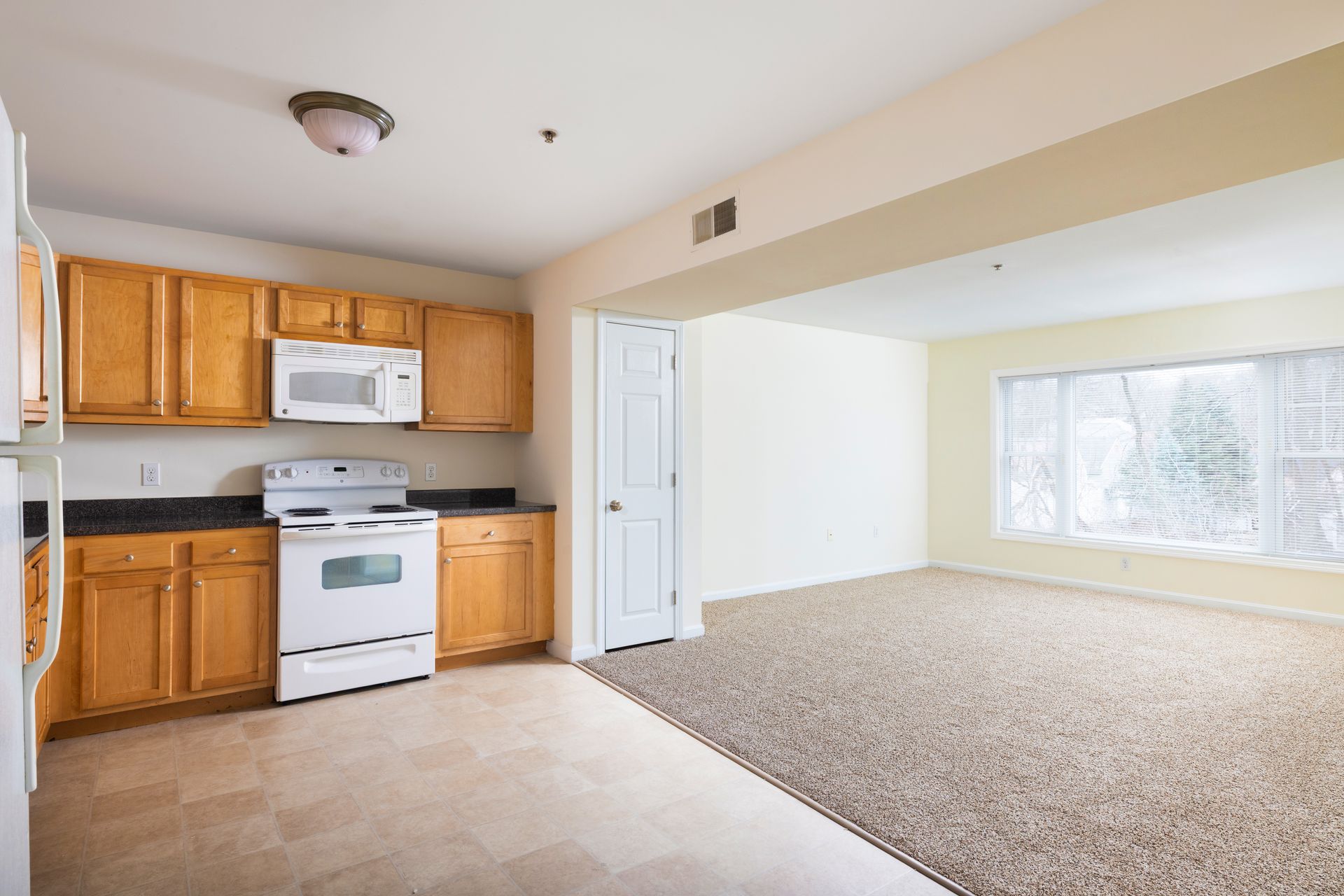 Empty kitchen and living area with wood cabinets, white appliances, and carpeted room by a large window