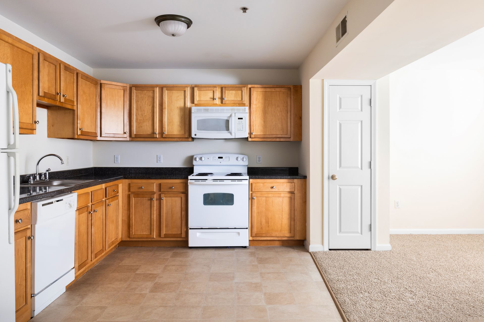 Small kitchen with wood cabinets, white appliances, and beige tile next to carpeted living area