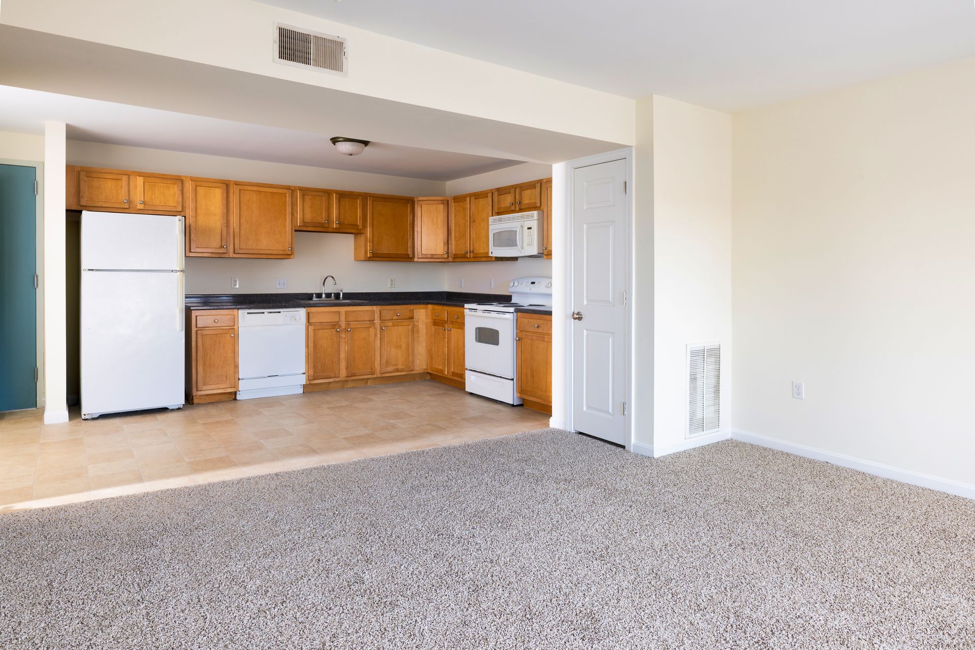 Empty living room with carpet and open kitchen, featuring white appliances and wood cabinets.