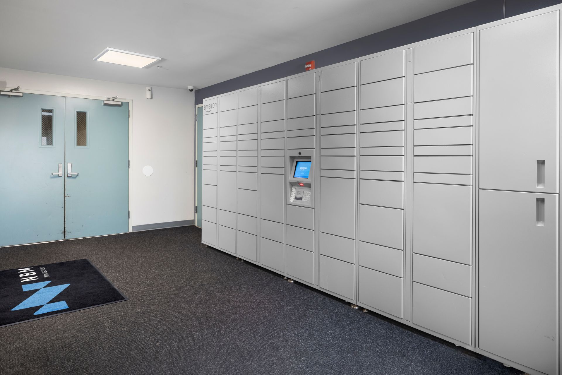 Lobby with gray parcel lockers, a keypad kiosk, blue double doors, and a black floor mat.