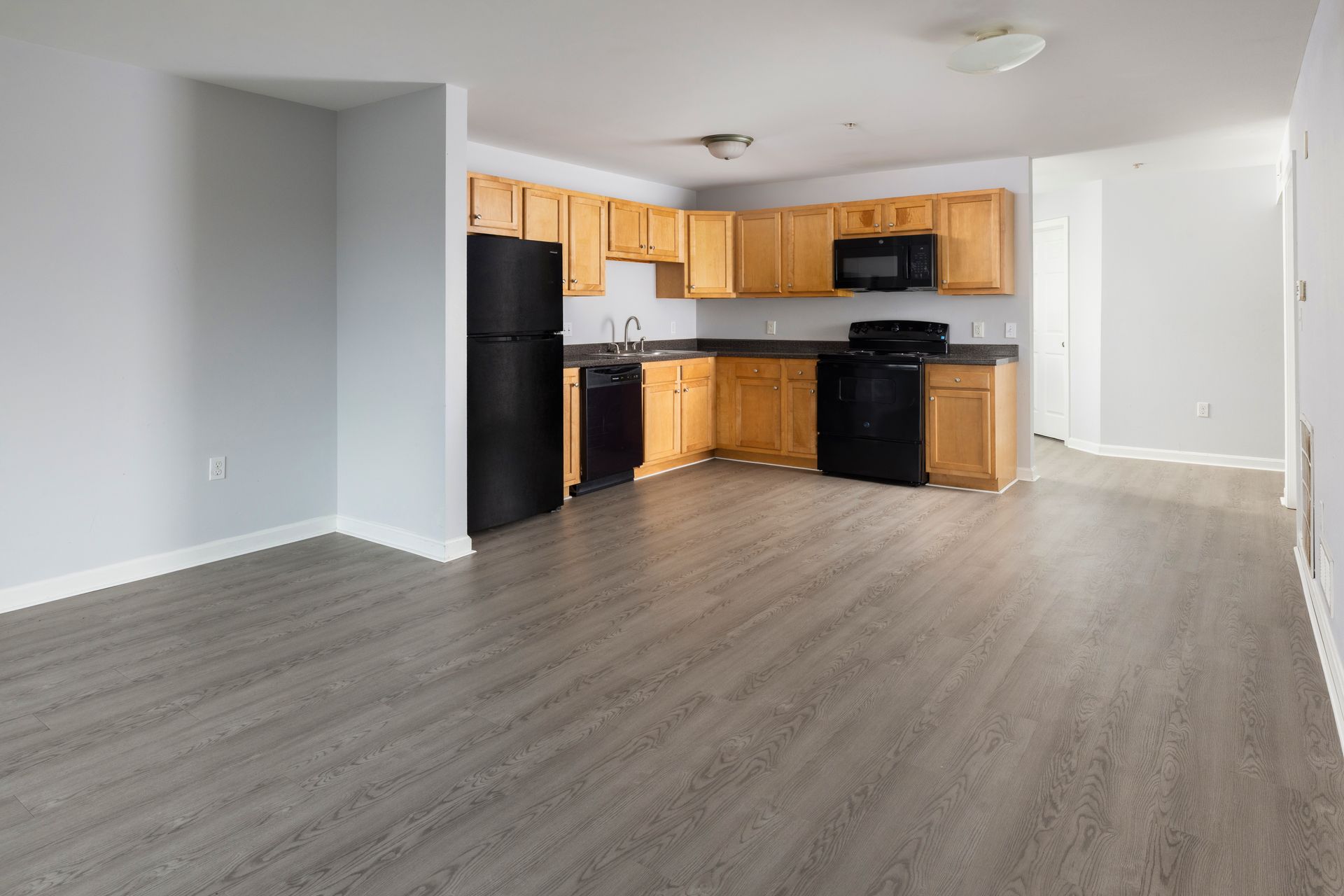 Empty open-plan kitchen and living area with black appliances, wood cabinets, and gray flooring