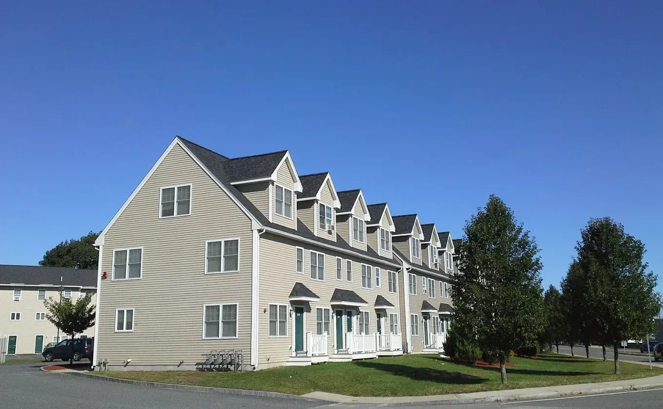 Three-story beige apartment building with white trim and dormer windows on a sunny street