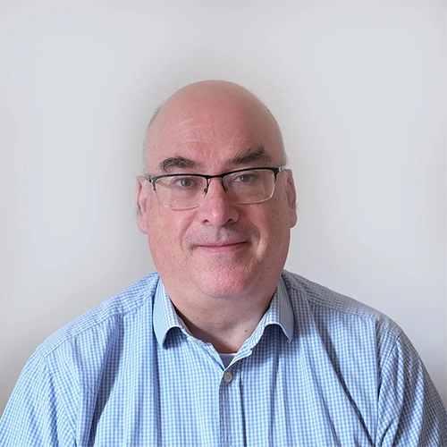 A bald man wearing glasses and a blue shirt is sitting in front of a white wall.
