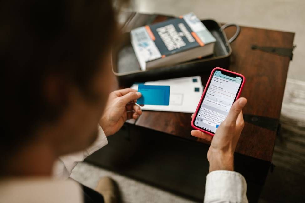 Person holding a smartphone, reviewing a mobile banking card and paper documents at a desk.