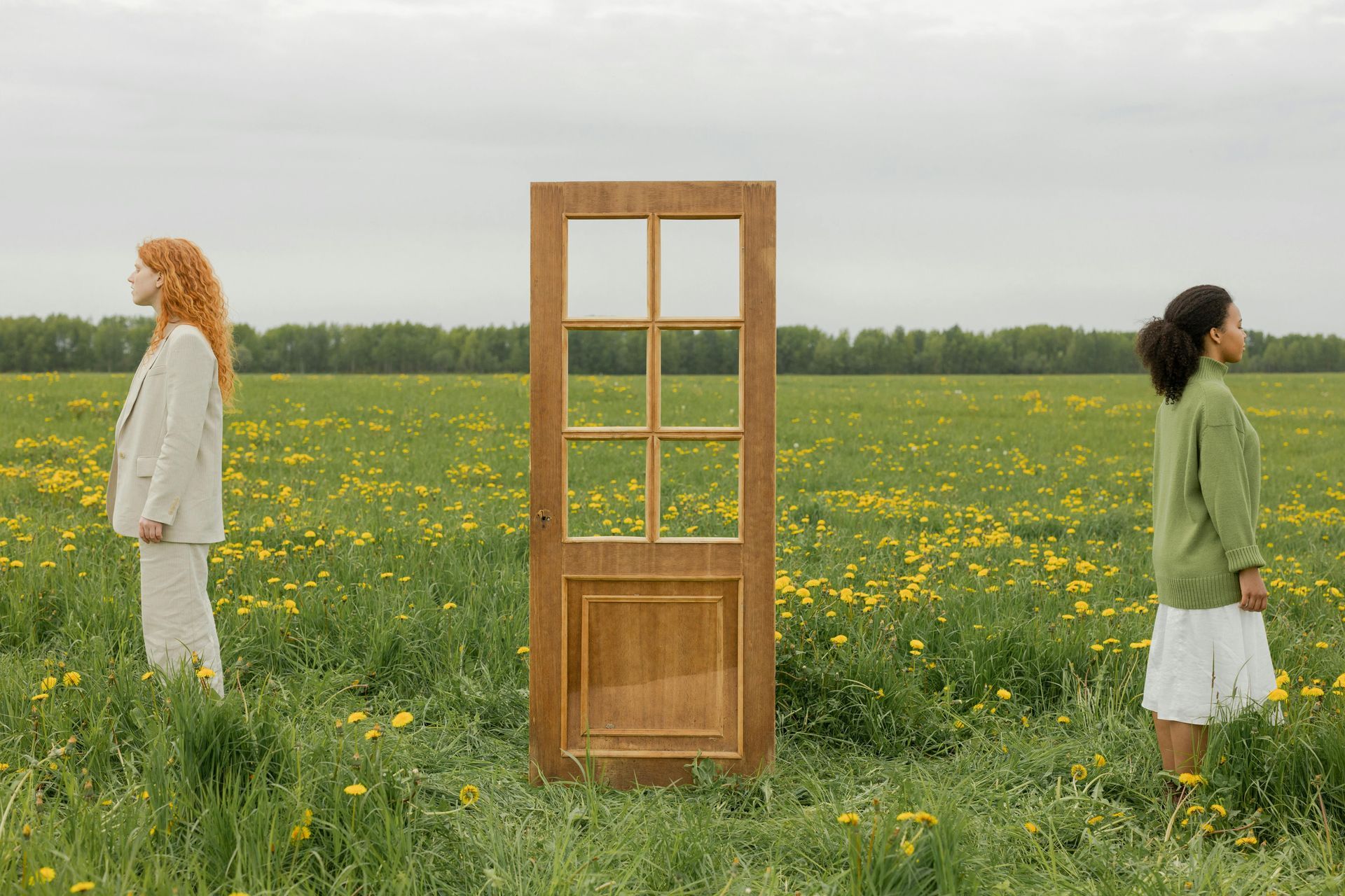 Twee vrouwen staan in een bloemenveld naast een houten deurkozijn onder een grijze hemel.